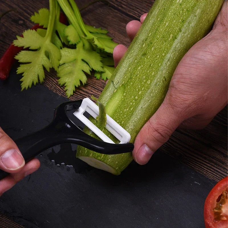 Hands use a black-handled ceramic peeler to peel a green zucchini on a dark cutting board, with celery and tomato nearby.