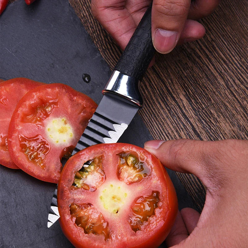 Ein gerilltes Küchenmesser schneidet eine reife Tomate auf einem dunklen Schiefer-Schneidebrett, daneben liegen Tomatenscheiben.