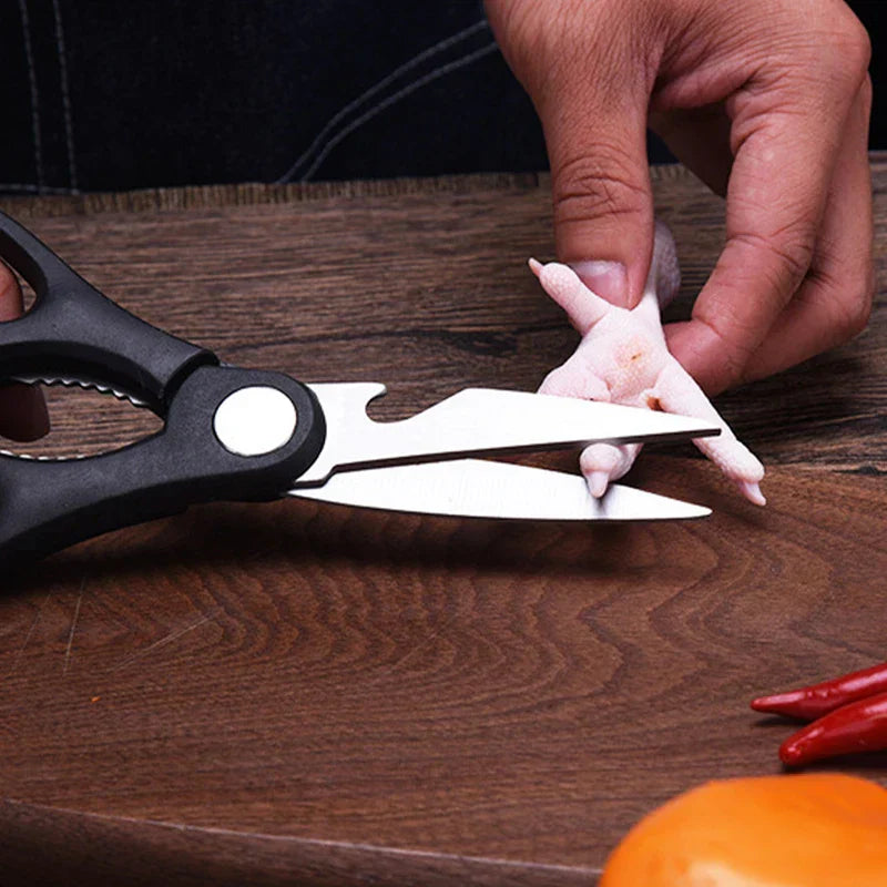 Hand holds a chicken foot on a wooden table while a pair of kitchen scissors cuts the chicken foot, with chili peppers and an orange nearby.