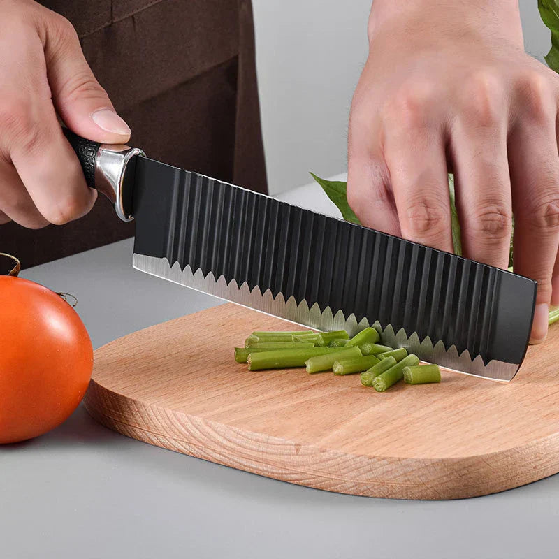 A person cuts green vegetable stalks on a wooden cutting board with a single black textured kitchen knife, a tomato sits beside the board.