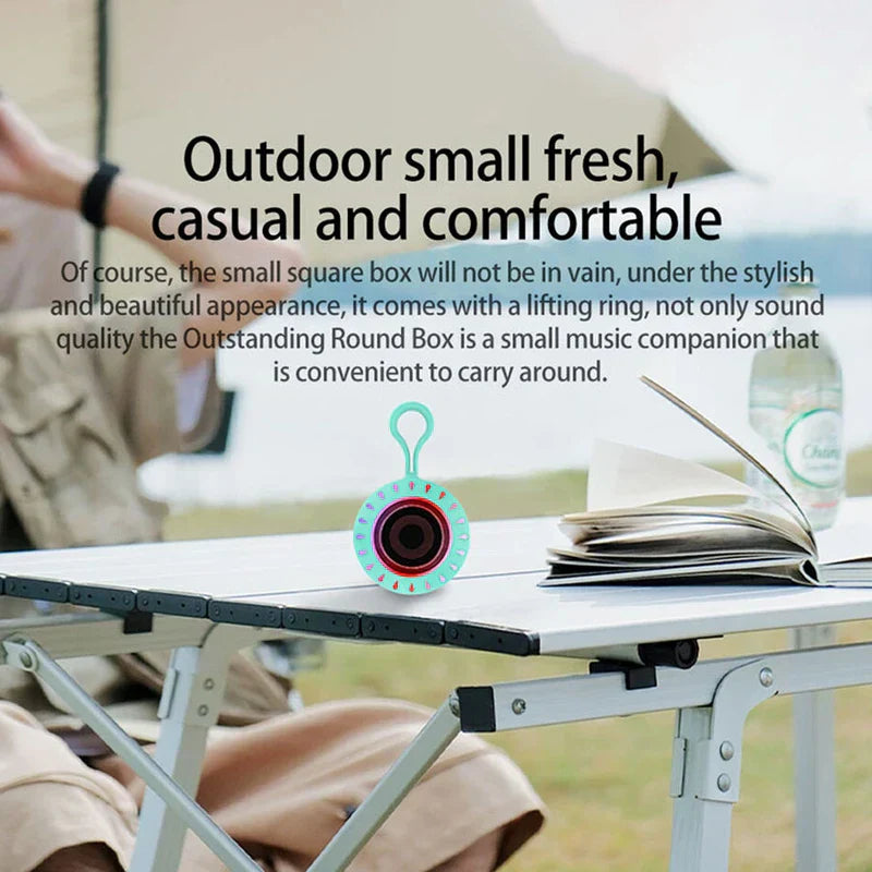 Portable Bluetooth Speaker - Small teal speaker with hanging loop placed on a folding camping table outdoors, with a person and open book visible in the background