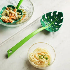 Strainer Spoon - Green monstera leaf utensil resting on a marble countertop next to bowls of cooked rotini and noodles