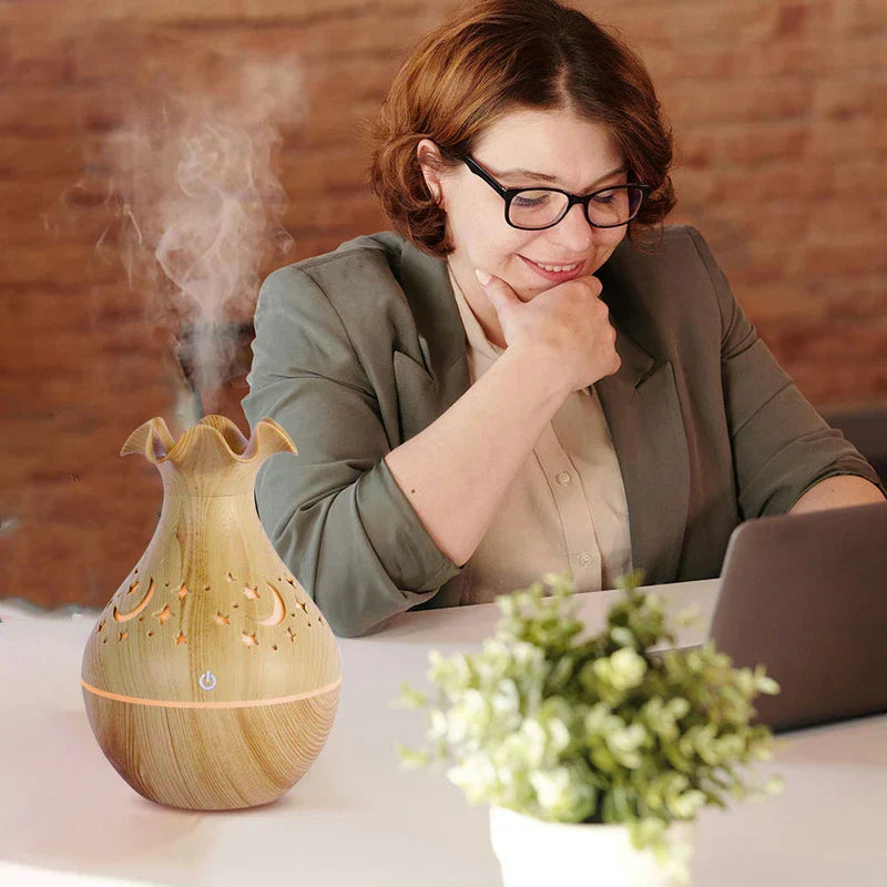 diffusor für ätherische öle - Diffusor für ätherische Öle - Vasenförmiger Diffusor mit Holztextur, der aus seiner ausgestellten Öffnung Nebel abgibt, mit warmem orangefarbenem Licht durch Ausschnitte, auf einem Schreibtisch neben einer sitzenden Person, die einen Laptop benutzt