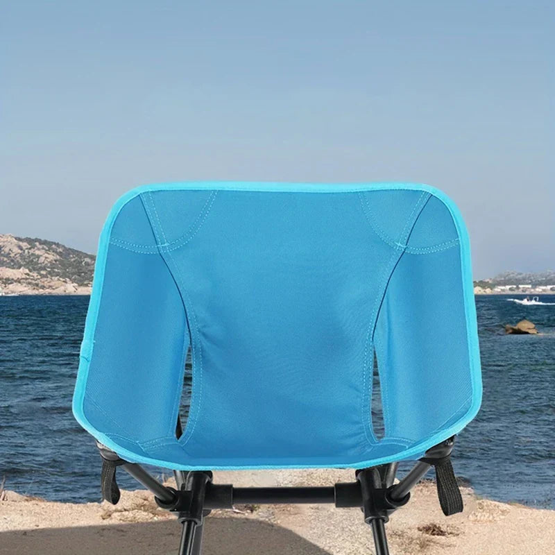 Folding Camping Chair - Back view of a bright blue folding camp chair set on sandy shore with ocean and coastal hills in the background