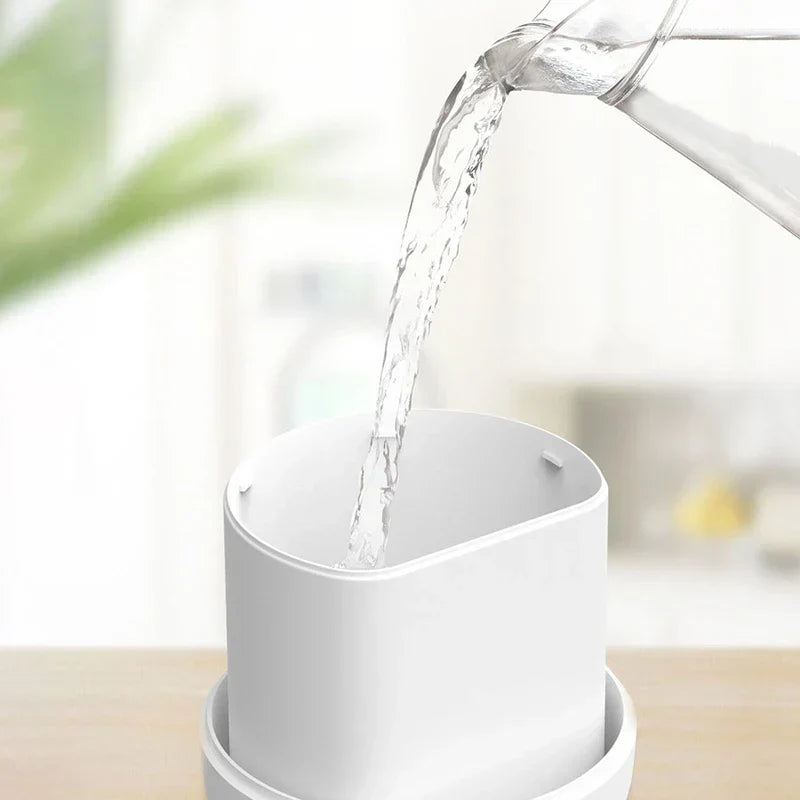 Humidifier Diffuser - Water being poured from a glass pitcher into a white cylindrical container on a light wood surface