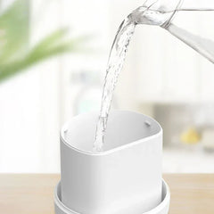 Humidifier Diffuser - Water being poured from a glass pitcher into a white cylindrical container on a light wood surface