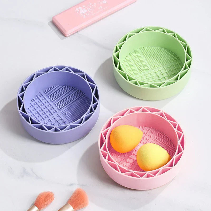Makeup Brush Cleaning Mat - Three round silicone bowls in purple, green and pink; pink bowl holds two makeup sponges, with makeup brushes and a pink case on white marble.