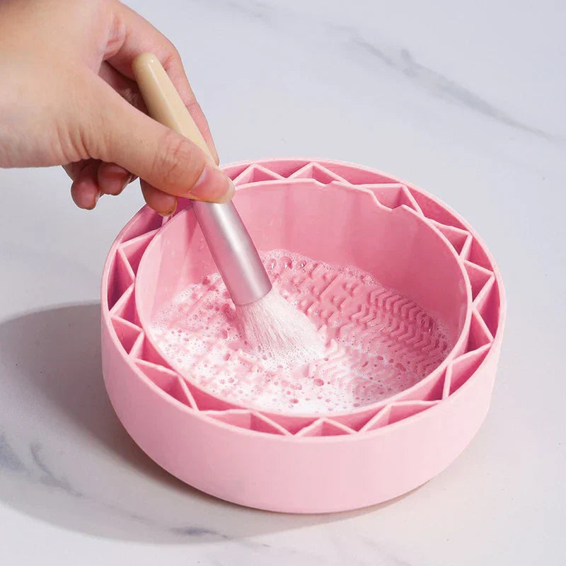 Makeup Brush Cleaning Mat - Hand holding a makeup brush, cleaning the brush head in soapy water inside a pink round silicone bowl.