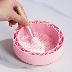 Makeup Brush Cleaning Mat - Hand holding a makeup brush, cleaning the brush head in soapy water inside a pink round silicone bowl.