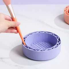 Makeup Brush Cleaning Mat - Light purple silicone makeup brush cleaning bowl on white surface, hand holding orange-handled makeup brush inserted into its edge slot