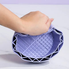 Makeup Brush Cleaning Mat - Hand squeezing a light purple flexible silicone cleaning bowl, showing its textured interior with multiple raised pattern zones