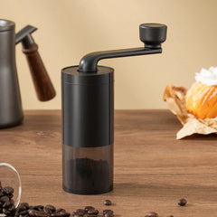 Coffee Bean Grinder - Black manual coffee grinder with ground coffee in its lower chamber, sat on a wooden surface alongside coffee beans