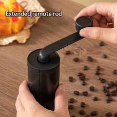 Coffee Bean Grinder - Two hands operating the grinder's extended crank handle, with loose whole coffee beans scattered on the wood table surface below