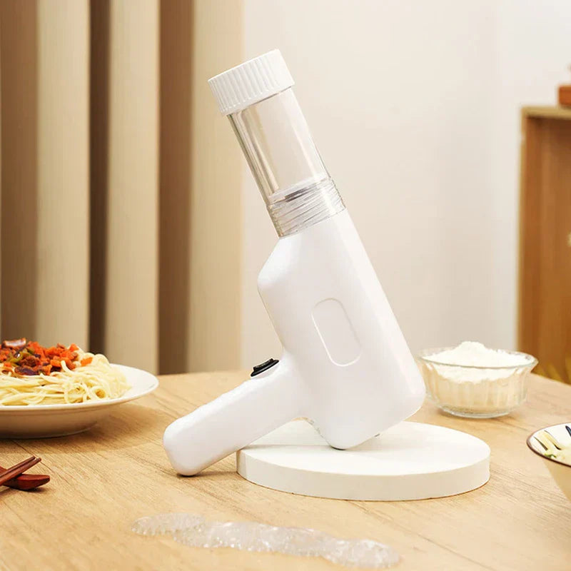 Manual Pasta Maker - White handheld kitchen tool resting on a round white display stand on a light wood table, with a plate of sauced noodles, small bowl of flour, chopsticks, and partial second dish placed nearby