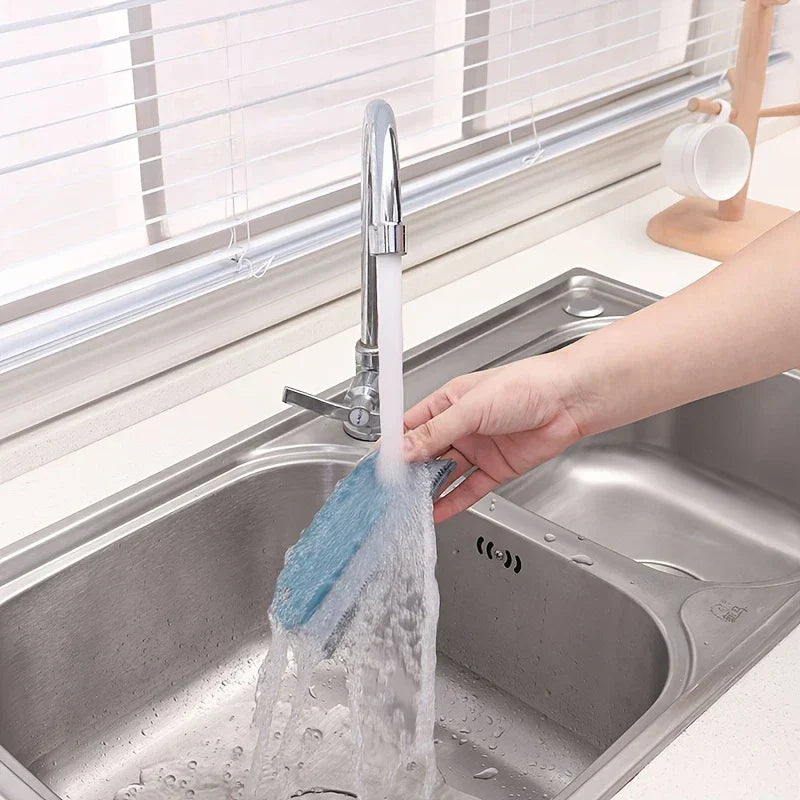 Dusting Brush - Hand holding a blue cleaning brush under running faucet water over a stainless steel double kitchen sink