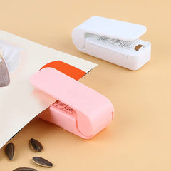 Heat Sealer - A pink mini bag sealer clipping a snack bag edge, with a white mini bag sealer placed nearby on a tan surface.