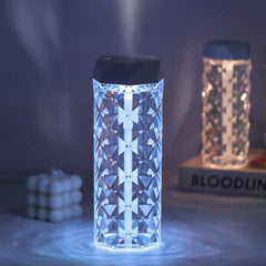 Cool Mist Humidifier - Illuminated humidifier emitting fine mist, with black top cap and faceted clear body, placed on a desk with decorative items in the background