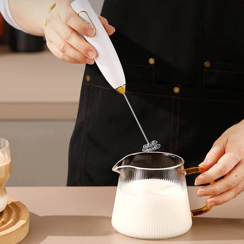 Electric Milk Frother - Person using a white frother to whip milk in a ribbed glass pitcher, holding the pitcher steady with their other hand