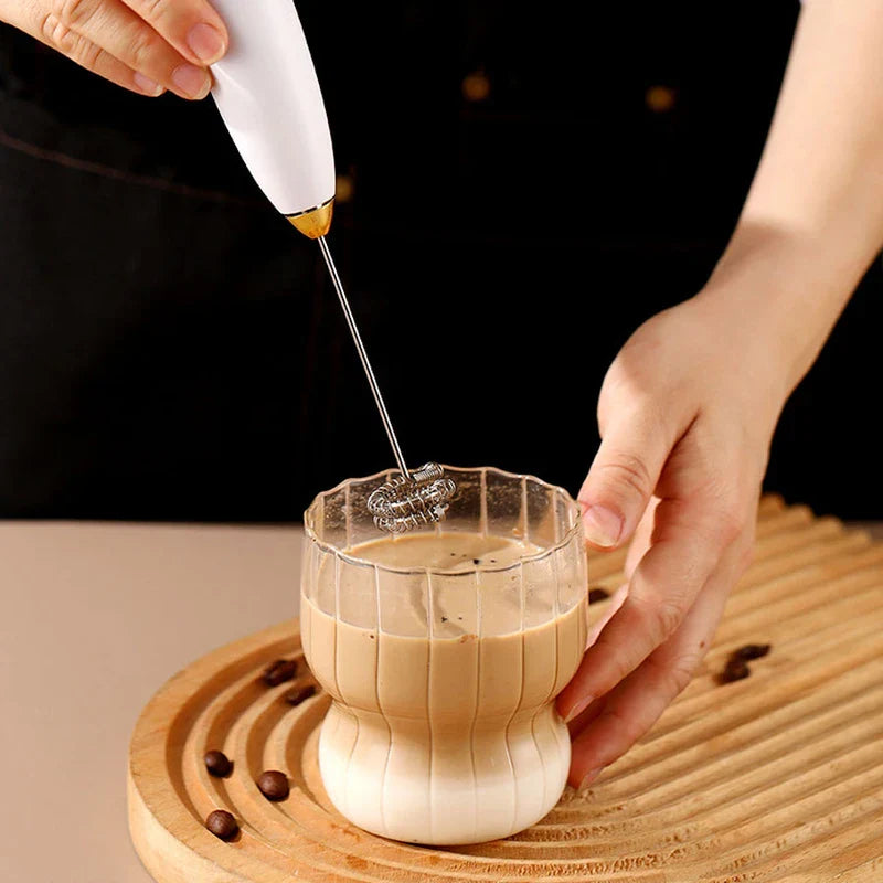 espumador de leche eléctrico - Mano sosteniendo un batidor eléctrico portátil mezclando una bebida de café en capas en un vaso acanalado, segunda mano estabilizando el vaso sobre una bandeja de madera con granos de café dispersos