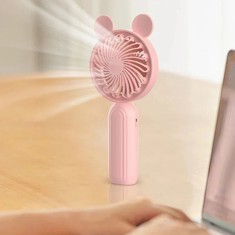 Mini Manual Handheld Squeeze Fan - Cute pink handheld fan with bear ears rests upright on a light wood desk, with a person’s hands typing on a laptop to its right.