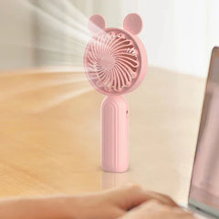 Mini Manual Handheld Squeeze Fan - Cute pink handheld fan with bear ears rests upright on a light wood desk, with a person’s hands typing on a laptop to its right.