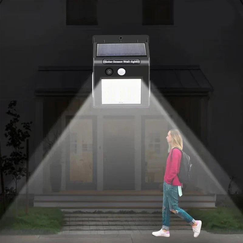 A person walking near a house at night is illuminated by an outdoor motion sensor light mounted on the home's exterior.