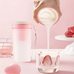 Pink accented portable juicer blender stands on a pink surface as a hand pours creamy liquid into a glass with strawberry slices, with strawberries nearby.