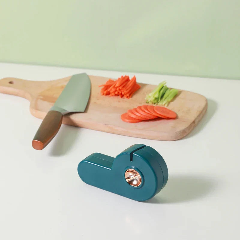 Manual Knife Sharpener - A teal sharpener sits on a white countertop in front of a wooden cutting board holding sliced vegetables and a knife