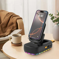 Car Phone Stand Holder - Black foldable phone stand speaker with RGB bottom lights, holding a smartphone, on a round light wood table next to a mug of coffee and a potted plant.