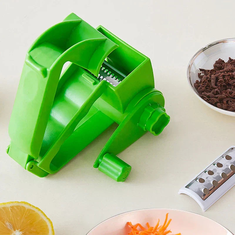 Vegetable Grater - Green rotary grater placed on a light countertop, with shredded carrot, chocolate shavings, a lemon half, and a spare blade nearby.