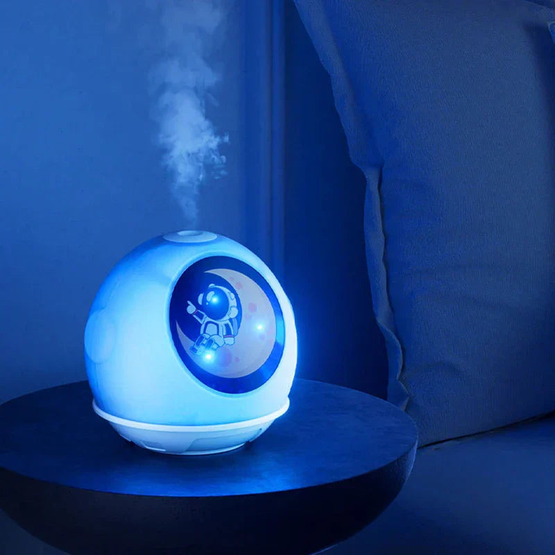 Space Theme Mist Maker - Round white humidifier with astronaut window panel emits mist, lit blue, sitting on a dark round side table