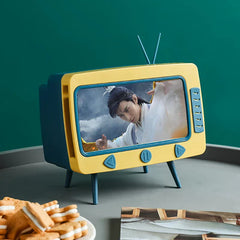 Yellow and teal retro TV-shaped novelty tissue dispenser playing a video, placed on a table with cookies and a magazine nearby