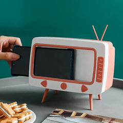 Hand inserting a phone into a white and coral retro TV-shaped novelty tissue dispenser, with cookies and a magazine on the table below