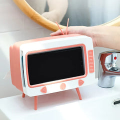 Pink and white novelty tissue dispenser shaped like a vintage TV, holding a smartphone on a bathroom sink counter.