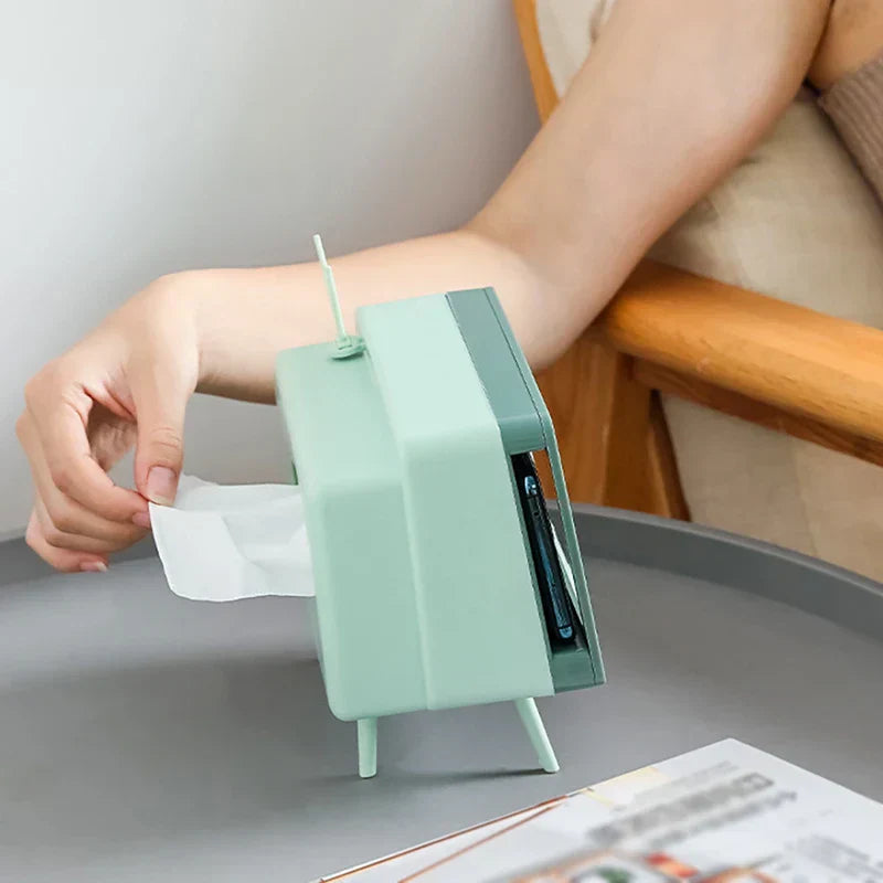 Light green novelty tissue dispenser in retro TV form, with a hand pulling a tissue from its side, on a round table.
