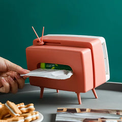 Side view of a pink novelty tissue dispenser shaped like a vintage TV, with a hand pulling a tissue from the rear opening, on a dark surface