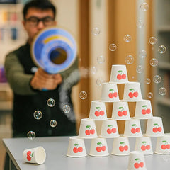 Person holds a blue and yellow bubble gun aimed at a stack of cherry-printed paper cups
