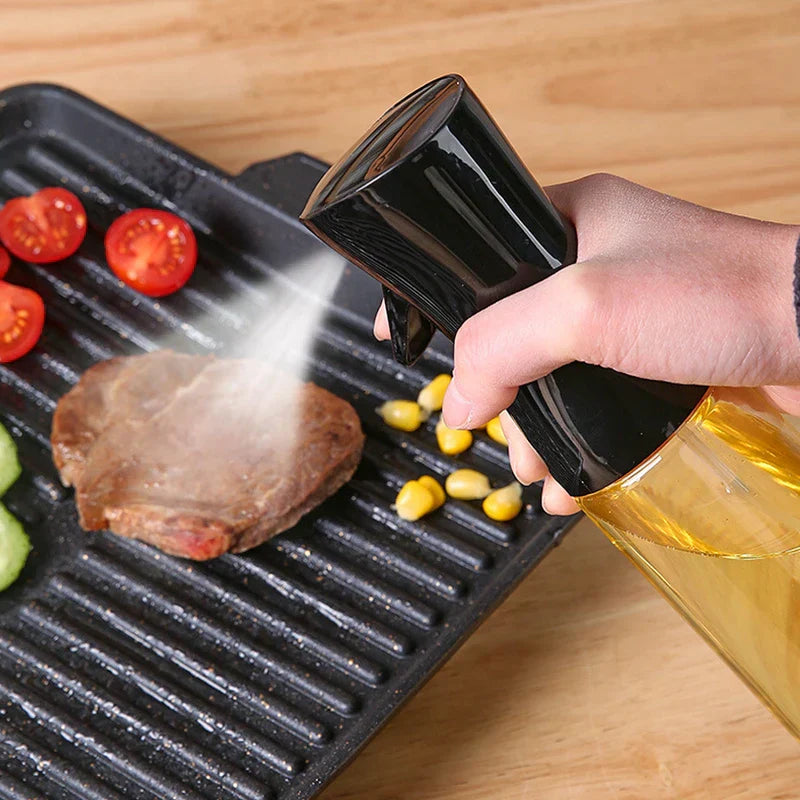 Oil Sprayer - Hand holding and depressing a black oil spray bottle, misting oil onto a steak on a ridged grill pan with tomato and corn pieces nearby
