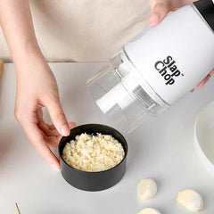 Garlic Mincer - User pouring freshly minced garlic from the chopper into a small black storage cup, with peeled garlic cloves on the countertop