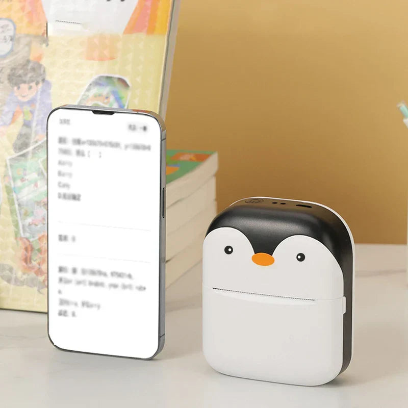 Impresora térmica de bolsillo - Impresora portátil con estilo de pingüino colocada junto a un teléfono inteligente que muestra una lista imprimible, con libros apilados al fondo