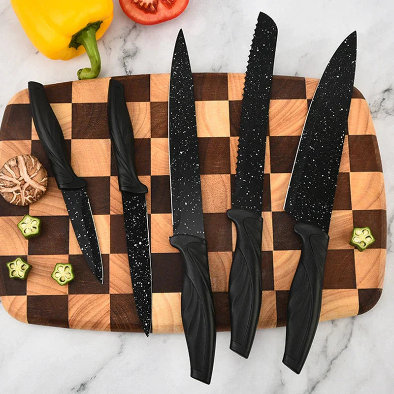 Five black speckled knives from a kitchen utensil set arranged on a checkered wooden cutting board with vegetables around it.