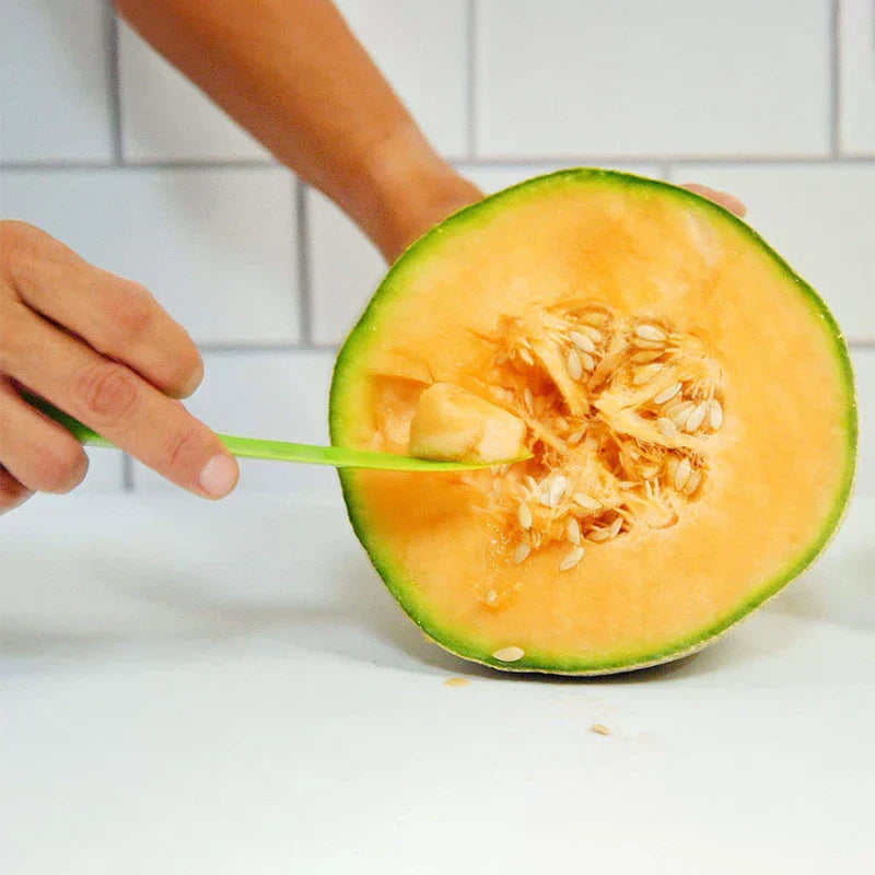Hand using green plastic melon slicer to scoop flesh from halved cantaloupe on white countertop