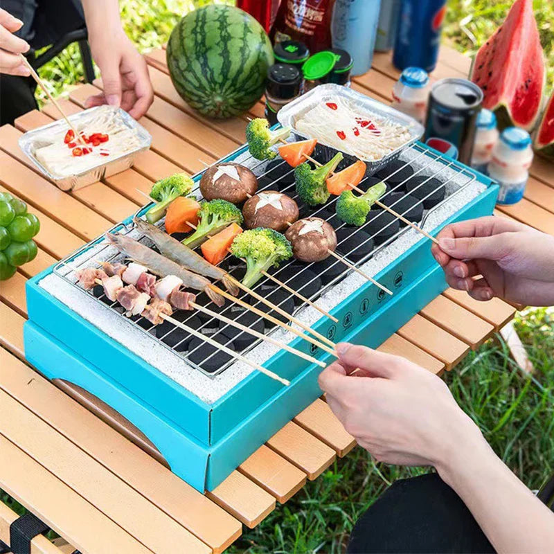 People using a teal disposable grill with a grill grate to cook shrimp, veggie, and meat skewers on an outdoor picnic table