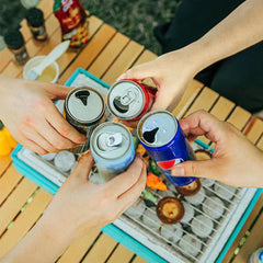 Four hands clinking beverage cans above a tabletop grill with grill grate holding cooking food