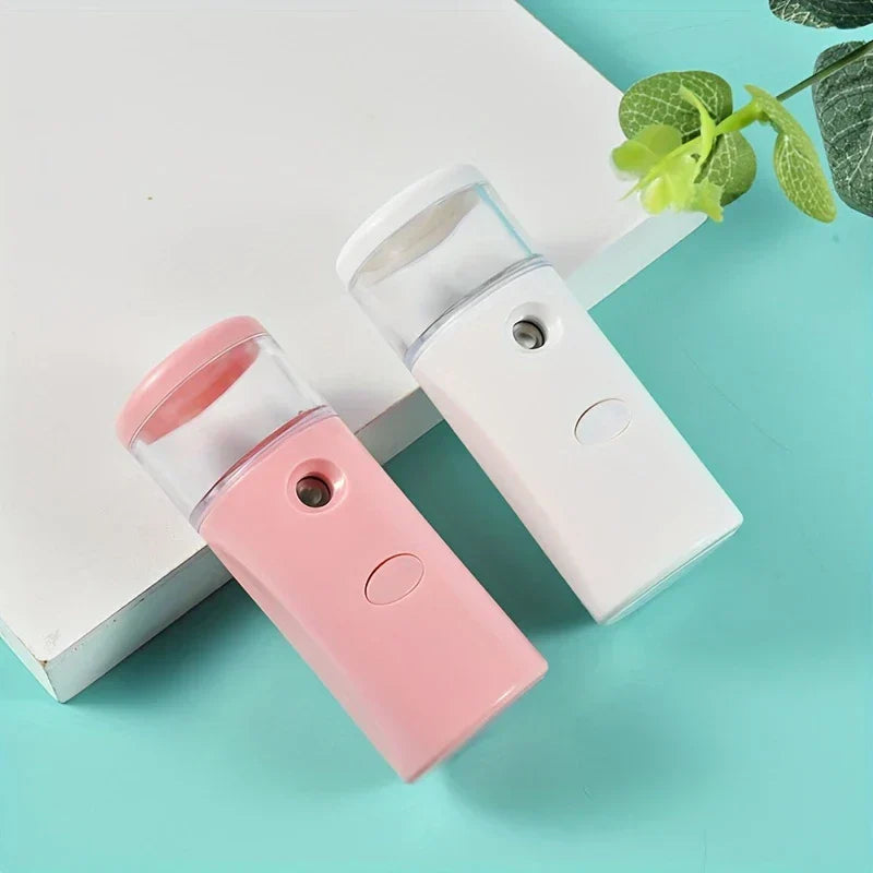 Portable Nano Facial Mist Sprayer - Pink and white portable nano facial mist sprayers resting on a white block over a teal surface with a leafy sprig