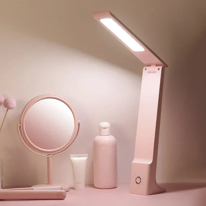 Pink led desk lamp lit on a pink tabletop alongside a pink mirror, skincare bottles, and pink decorative items against a light neutral wall