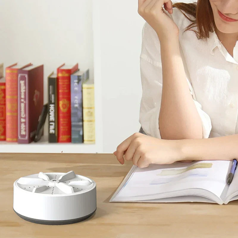 Compact grey and white mini washing machine sits on wooden desk beside open notebook, with bookshelf and person in background