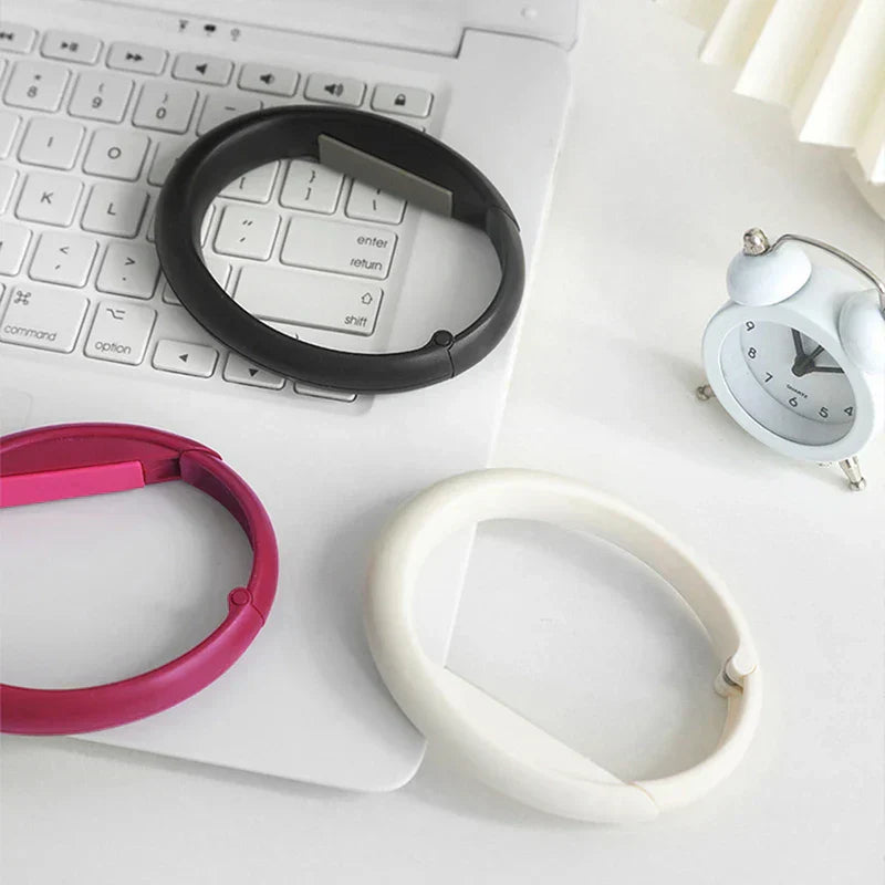 Handtaschenhaken - Drei Handtaschenhaken in Schwarz, Pink und Weiß liegen auf einer weißen Laptop-Tastatur neben einem kleinen Wecker