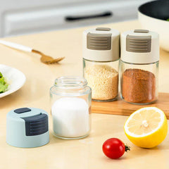 Salt Shaker Bottle - Multiple salt shaker bottles (one disassembled) on a counter with lemon, cherry tomato, and spice-filled jars