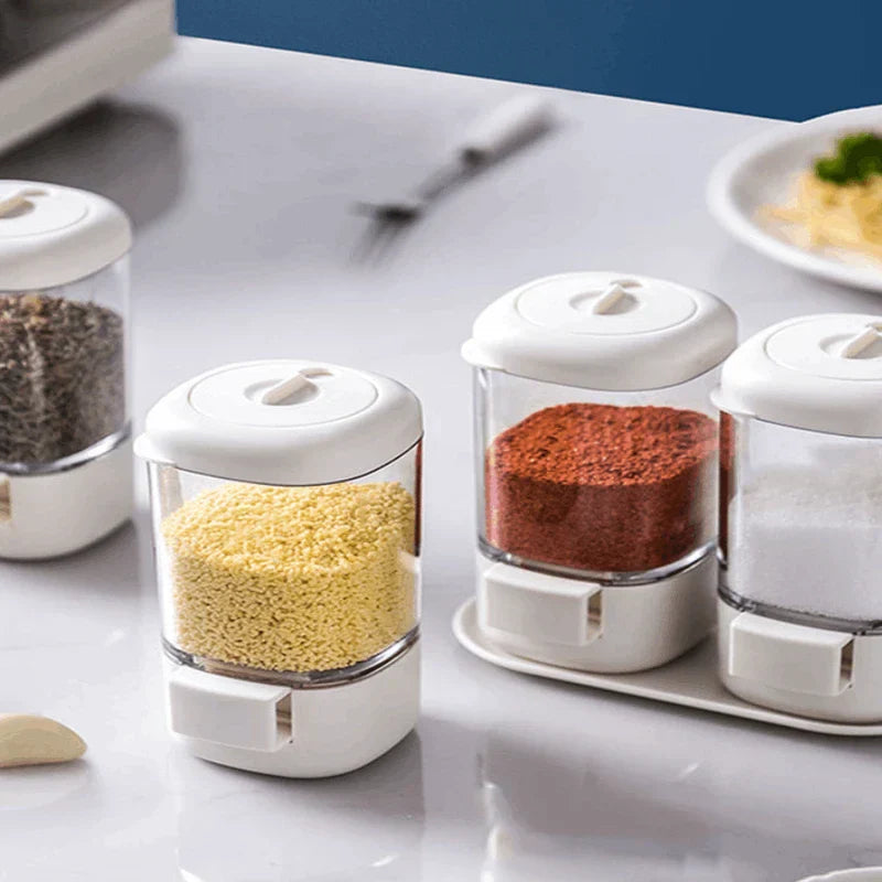 Frascos de especias transparentes con botón pulsador y tapas blancas que contienen diferentes condimentos en una encimera de cocina